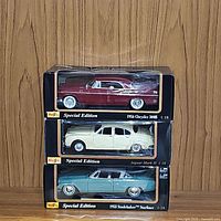 Three Maisto Special Edition 1:18 diecast model cars stacked in their original boxes with clear packaging: red 1956 Chrysler 300B, cream 1959 Jaguar Mark II, light blue 1953 Studebaker Starliner.