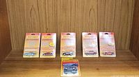 Seven Matchbox vehicles in original sealed blister card packaging arranged in two rows on a wooden shelf. Six cars are displayed standing in blister packs, one pack lower centered.