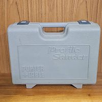Gray hard plastic carrying case with molded handle and latches, labeled 'Porter Cable Profile Sander' embossed on the front.