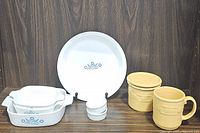 CorningWare Blue Cornflower dishes including casserole dishes and pie plate, white ceramic ramekins, and Longaberger butternut yellow mugs and lid arranged on wooden shelf.