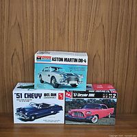 Front view of three model car kits: AMT 1951 Chevy Bel Air Convertible (blue and white), AMT 1957 Chrysler 300C (red), and Monogram Aston Martin DB-4 (silver) boxed kits stacked against wood background.