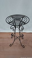 Brown metal plant stand with spiral circular top and scrollwork legs photographed against a neutral background.