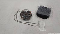 Round metal SAJAI bag decorated with various colored small stones, metal chain strap, black pouch wrapped in plastic, and an information card about the bag.