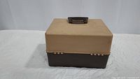 Closed two-tone fishing box with beige top and dark brown bottom, black handle on top.