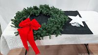 Table display of a green artificial Christmas wreath with a large red bow, and a white blow mold star with attached electrical cord for lights, both laid on a white table with black cloth underneath.