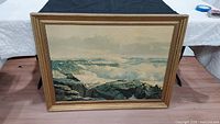Full view of the large framed ocean painting showing the ocean waves crashing on rocks with a cloudy sky above.