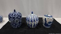 Three blue and white lidded ginger jars of varying sizes shown together on black cloth.