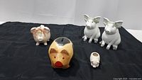 Five pig-themed items displayed on black cloth: two white ceramic piggy banks with green wreaths and silver hooves, one pink piggy bank with floral wreath, one wooden piggy bank with red mouth, and one small white toothpick holder shaped like a pig.