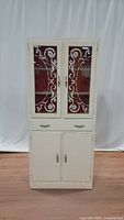 Front view of vintage white wooden kitchen cabinet with two glass doors featuring decorative floral and scroll designs.