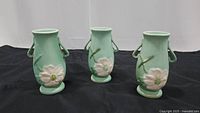 Three green Weller vases on black cloth, showing front floral decoration and side handles.