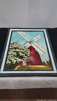 Framed painting showing full image of red windmill with white blades and greenery below.