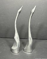 Two tall polished aluminum swan sculptures with elongated necks, standing against a black background.