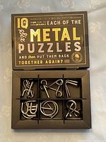 Photo of the opened box showing six separate metal wire puzzles in a black plastic tray in original packaging. Puzzle shapes vary with loops, bends, and interlocking components.