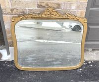 Front view of the gilt framed mirror leaning against brick wall, showing overall shape and detailing.