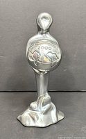 Front view of polished metal sculpture with globe marked 2000 held by a stylized female figure on organic base.