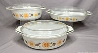 Set of three vintage Pyrex round casserole dishes with glass lids, displaying orange and brown geometric floral pattern on white glass.