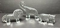 Three clear glass animal sculptures: lion, elephant, hippopotamus displayed on dark surface