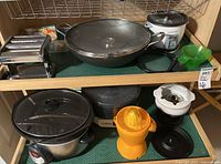 Shelf with cast iron wok, rice cooker, green manual juicer, black slow cooker with glass lid, yellow manual juicer, fondue pot with lid, and dehydrator.