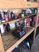 Wide view of wooden shelves holding assorted chemical bottles and a Schumacher battery charger with clamps on the lower shelf.