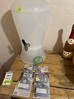 Wide view of white plastic beverage container with tap, showing wine bottle tags in front and Steam Whistle bottle opener on base.