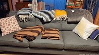 Three Mexican style blankets draped over a couch with two brown striped blankets and one gray blanket arranged on the couch cushions, two decorative pillows on the couch.