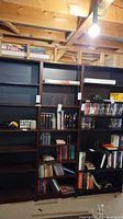 Three IKEA Billy bookcases in black-brown standing side by side filled with books and a few board games, in a basement with unfinished ceilings.