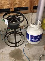 Metal burner and propane tank side-by-side on concrete floor in garage.
