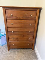 Front view of the wooden 5-drawer dresser with silver knobs against a beige wall and carpeted floor.