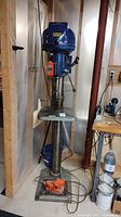 Front view of heavy-duty floor drill press with blue body, control levers, and attached adjustable worktable.