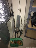 Three fishing rods with reels standing upright against a wall with a green tackle box containing lures in front.