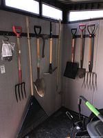Photo showing all ten garden tools hung on wall hooks in corner of room including pitchfork, shovel, rake, axe, leaf shovel, and packaging with small hooks.