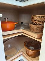 Corner cabinet shelf showing orange Lagostina pot with lid, clear glass bowls, and assorted wicker baskets stacked.