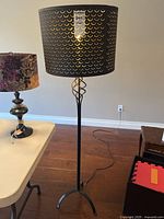 Full view of the floor lamp showing the black metal stand, decorative black perforated shade with gold interior, and tripod base on hardwood floor.