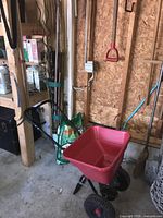 Full view of the red Earthway EV-N-SPRED spreader with the handle and wheels, a green bag of Scotts Turf Builder lawn food, and garden stakes leaning against the wall.