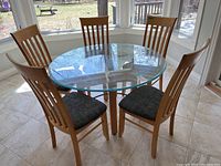 Full view of round glass top table with five wooden cushioned chairs around it, set near windows.
