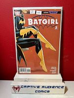 Photo of Batgirl #2 comic book in protective sleeve against red background with comic store branded stand.