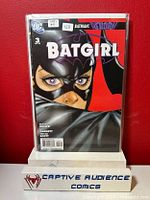 Batgirl #3 comic book sealed in protective plastic sleeve held upright against a red background with a small stand featuring the seller's logo.