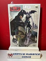 Photo of Batman #1 comic book in protective sleeve standing on display with Captive Audience Comics sign beneath it.
