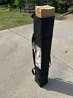 Photo of the black carrying bag with a box inside, standing upright on a concrete driveway with some grass and outdoor furniture in the background.