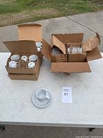 Two open cardboard boxes containing multiple white cups and matching saucers arranged inside.