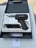 Full view of a black Archer soldering iron with a red trigger, metal solder tip, and coiled power cord inside a black plastic carrying case with white foam insert.