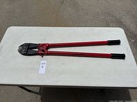 Full view of 36-inch bolt cutters placed on a white folding table showing red handles with black grips and black cutting jaws.