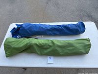 Two folded camping chairs in fabric carrying bags, one blue and one green, placed on pavement surface.