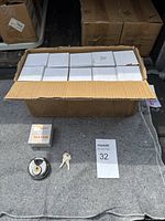 Opened cardboard box showing multiple smaller white boxes arranged inside, each presumably containing one disc lock and matching keys. One disc lock and two keys displayed outside the box with a white card numbered 32 nearby.