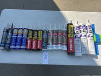 Full view of all 17 tubes of caulk and adhesives on a white table outdoors.