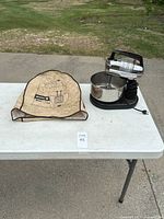 Photo of electric stand mixer with stainless steel mixing bowl and beige cloth cover on a white table outdoors.