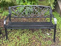 Full front view of the black metal garden bench placed outdoors on a patch of grass with greenery in the background. Shows decorative backrest and slatted seat clearly.