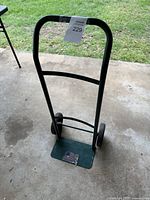Front view of black metal hand truck with small base platform and two wheels on concrete floor with grass background.