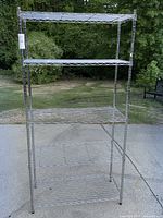 Full view of metal wire shelving unit placed outdoors on concrete with greenery in background.