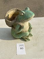 Side angle showing the seated ceramic frog holding a textured basket on its back, green and cream colors visible.